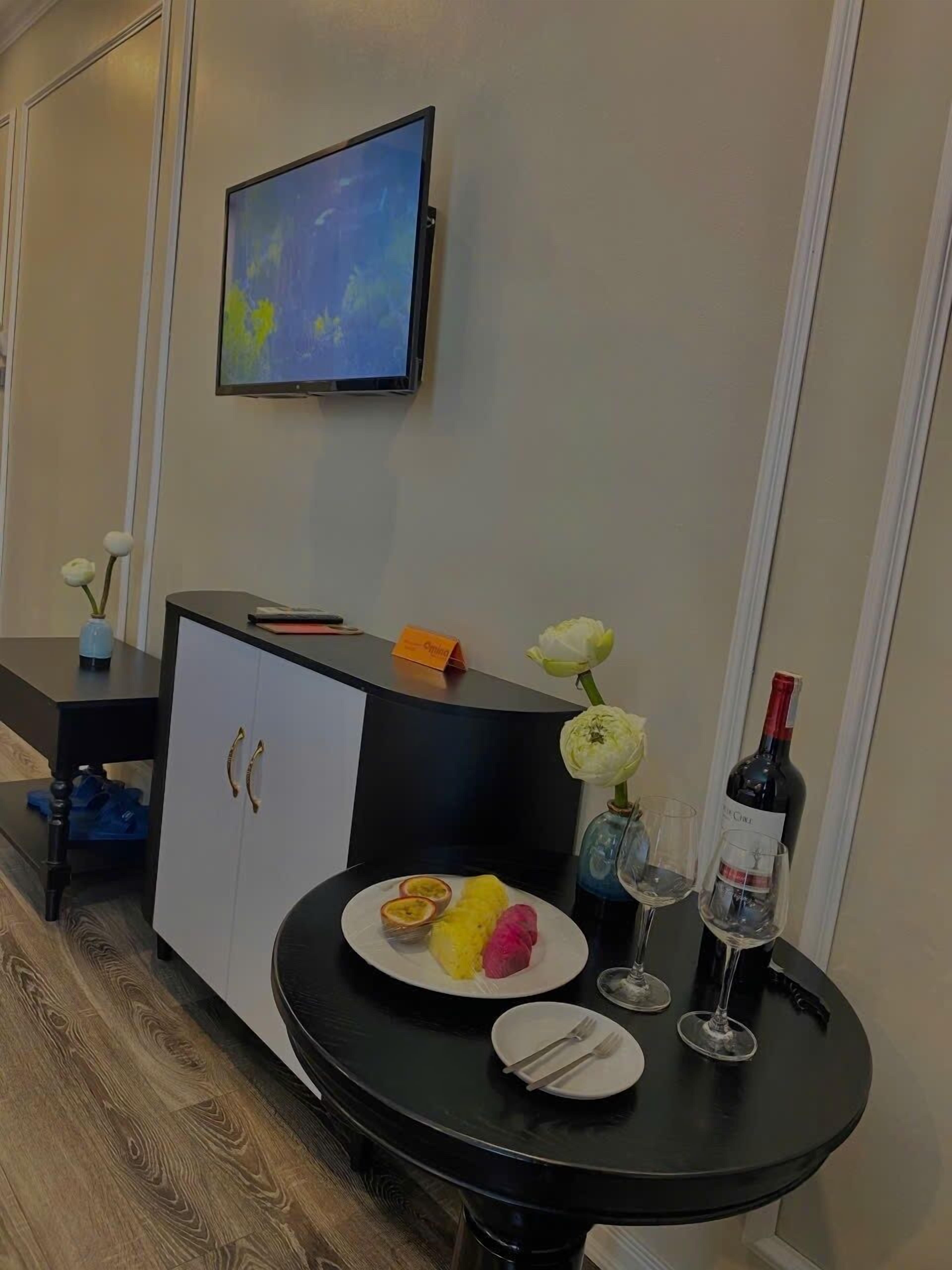 A hotel room with a black dining table set for two, a bottle of wine, and a vase with white flowers.