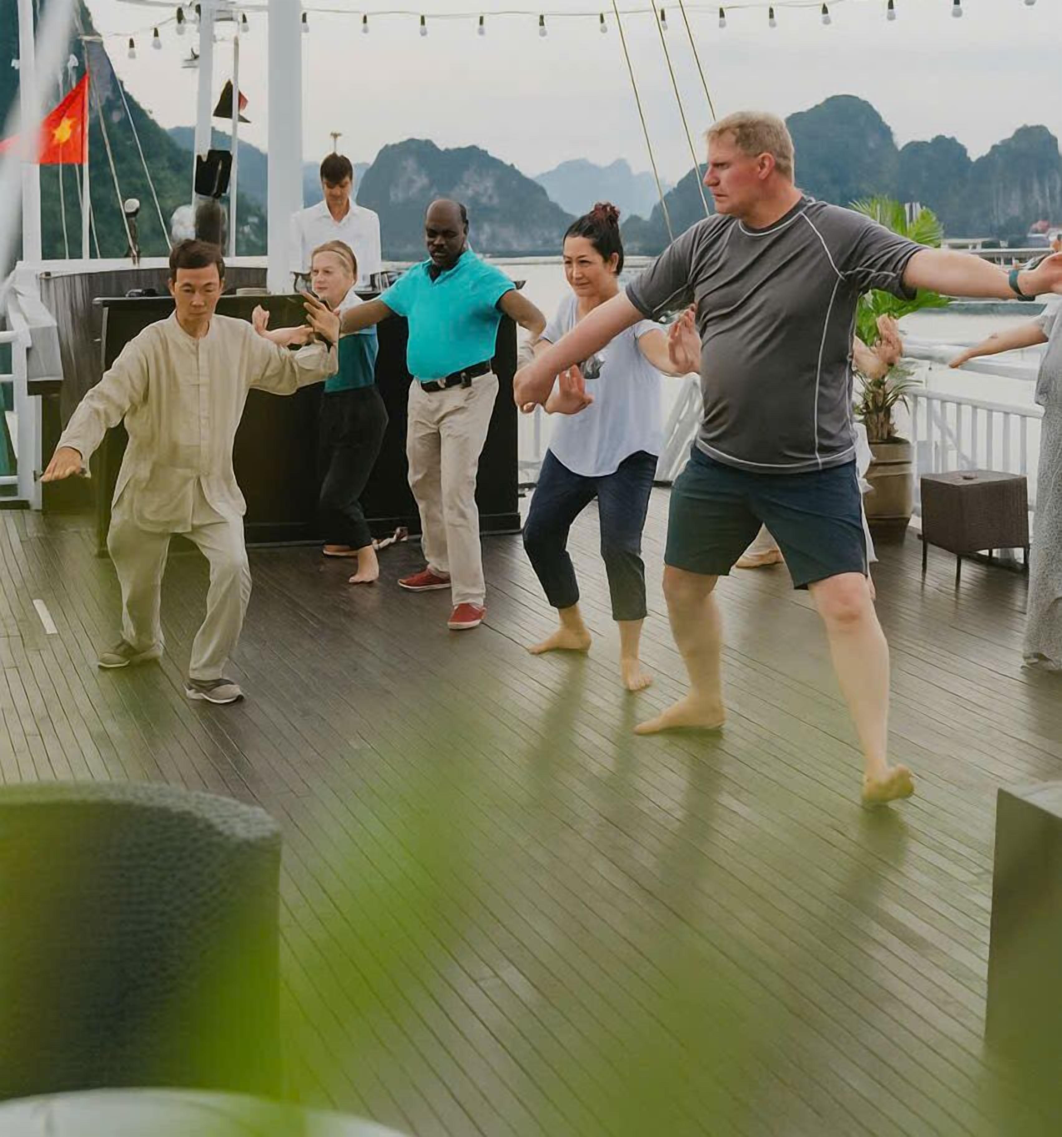 A group of people practicing Tai Chi on a wooden deck with a view of mountains and a flag in the background.