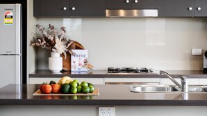 A modern kitchen with a stainless steel refrigerator, a gas stove, and a sink. There is a fruit bowl with tomatoes, limes, and avocados on the counter. A vase with dried flowers and a magazine titled "HOME" are on the shelf. - Villa Sangiovese - Unit One - 1-2 Heath Street (Evans Head)