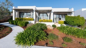 Une maison moderne à deux étages, avec une façade blanche, de grandes fenêtres et un jardin bien entretenu, agrémenté de diverses plantes et arbustes.