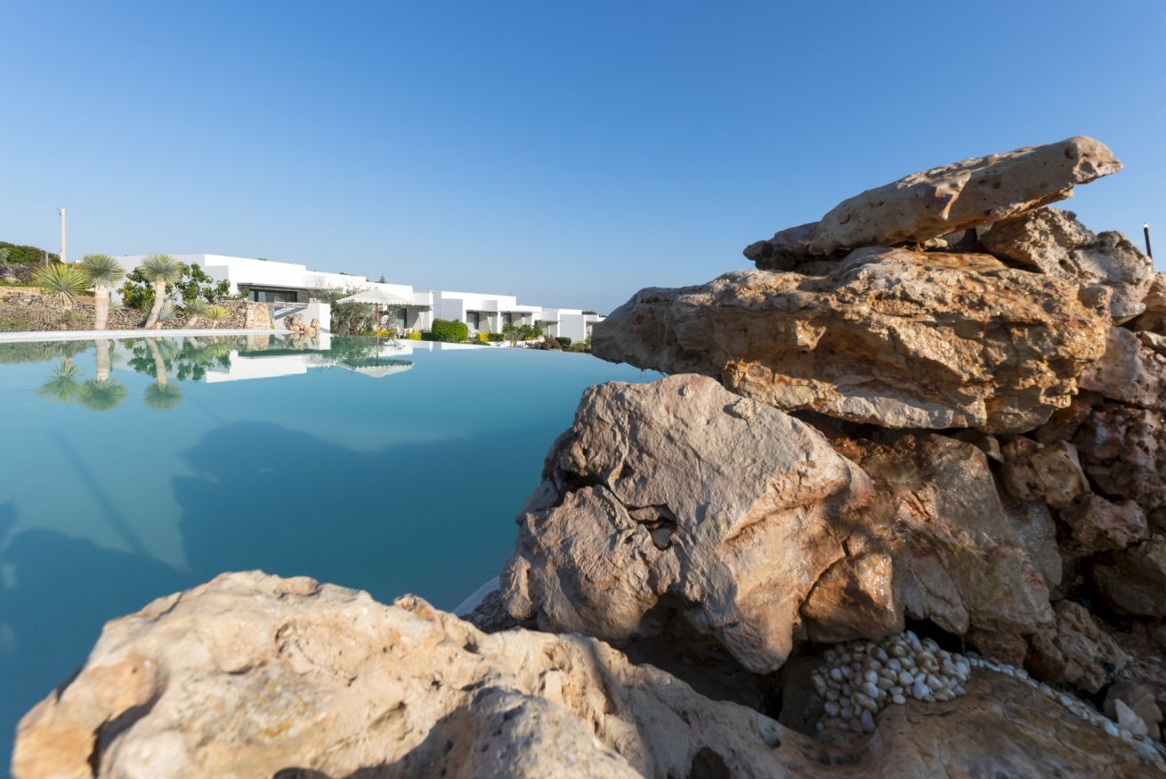 Une piscine à l’eau d’un bleu cristallin, entourée d’un paysage rocheux et bordée de bâtiments blancs en arrière-plan.