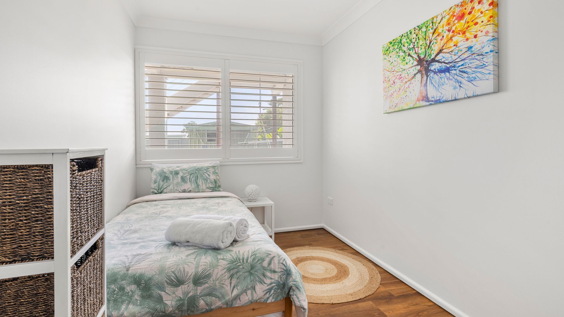 Een slaapkamer met een bed, een raam met zonwering, een schilderij aan de muur en een gevlochten mand.