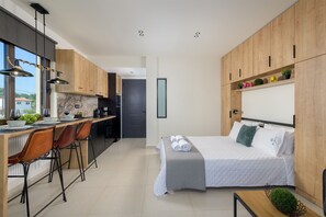 Ein modernes Hotelzimmer mit Küchenbereich, Holzschränken, einem Bett mit weißer Bettwäsche und einem kleinen Tisch mit einer Pflanze.