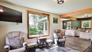 A spacious living room with a large window, a flat-screen TV, and a kitchen in the background. - A pet-friendly, creek side ranch house w/starlink (Port Angeles)