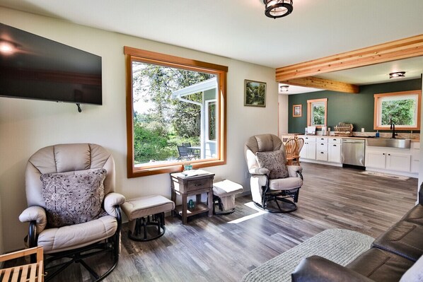 A spacious living room with a large window, a flat-screen TV, and a kitchen in the background.