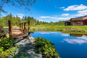 Eine Holzbrücke über einem stillen Teich, umgeben von viel Grün und einem roten Gebäude im Hintergrund.
