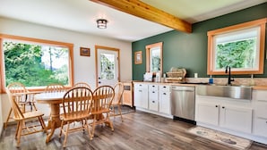A kitchen with wooden flooring, white cabinets, a dining table with chairs, and large windows with views of greenery. - A pet-friendly, creek side ranch house w/starlink (Port Angeles)