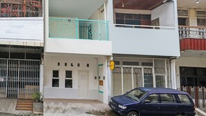Un edificio de dos pisos con fachada blanca, un balcón con barandillas turquesas y un coche azul estacionado adelante.