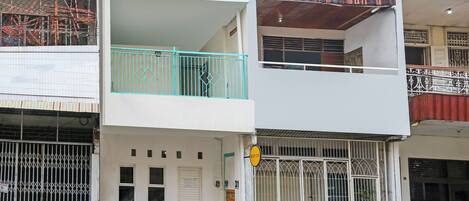 A two-story building with a white facade, a balcony with turquoise railings, and a parked blue car in front.