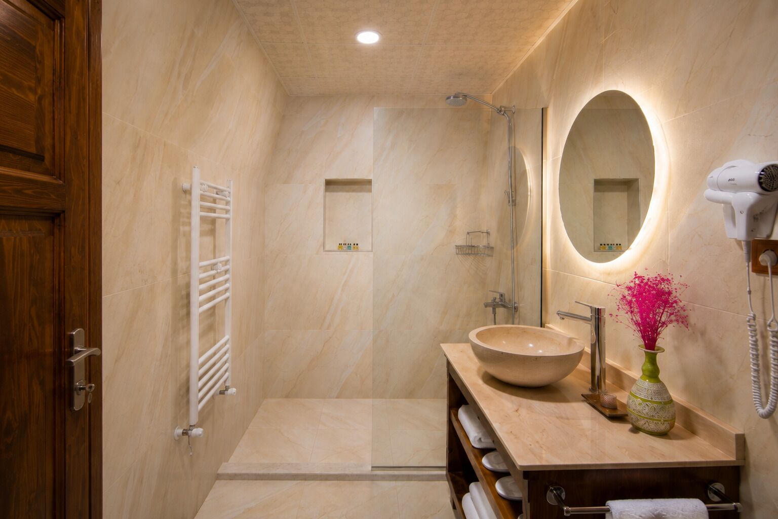 A modern bathroom with a large oval mirror, a beige sink, and a hairdryer on the wall.