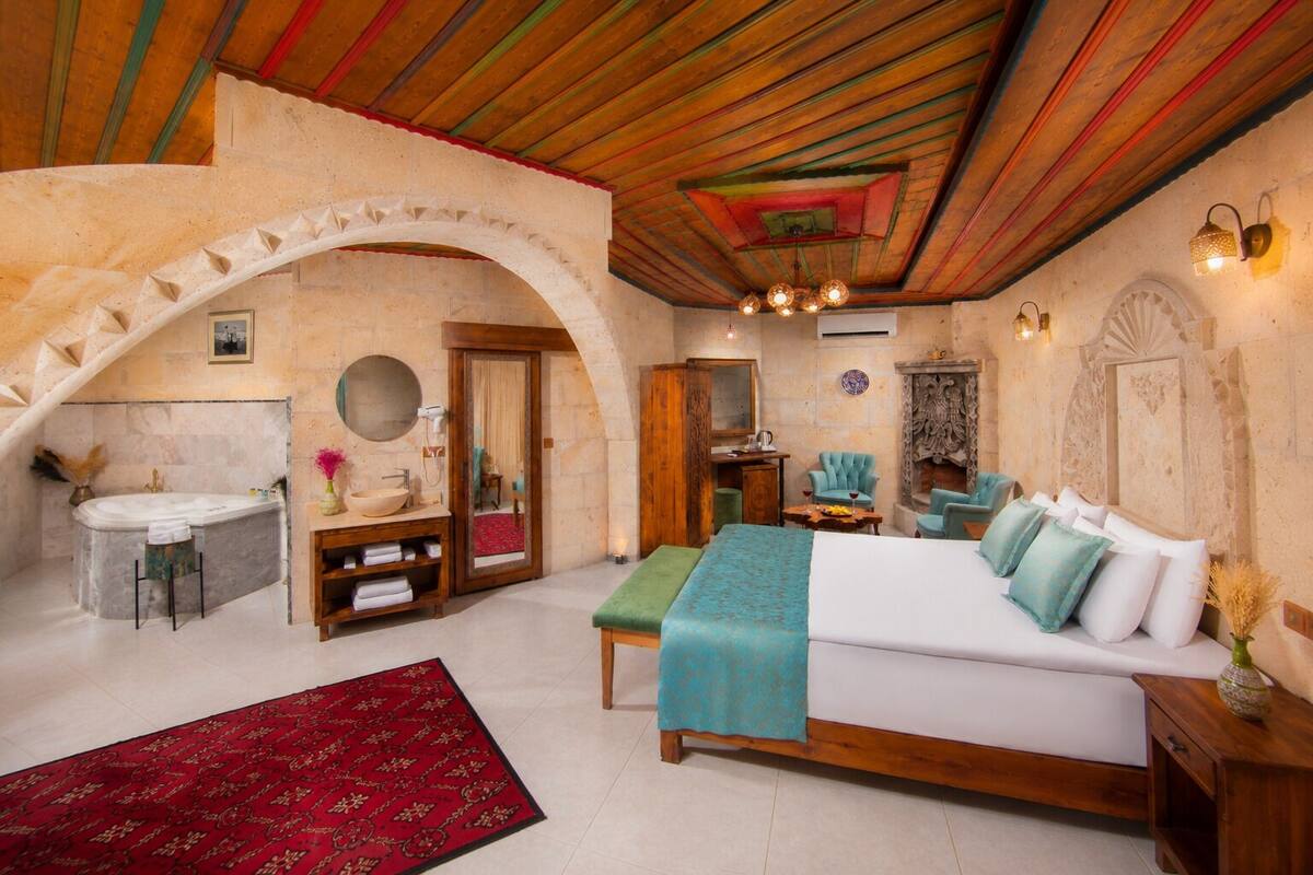 A bedroom with a large bed, a wooden bench, a vanity with a mirror, a fireplace, and a stone archway.