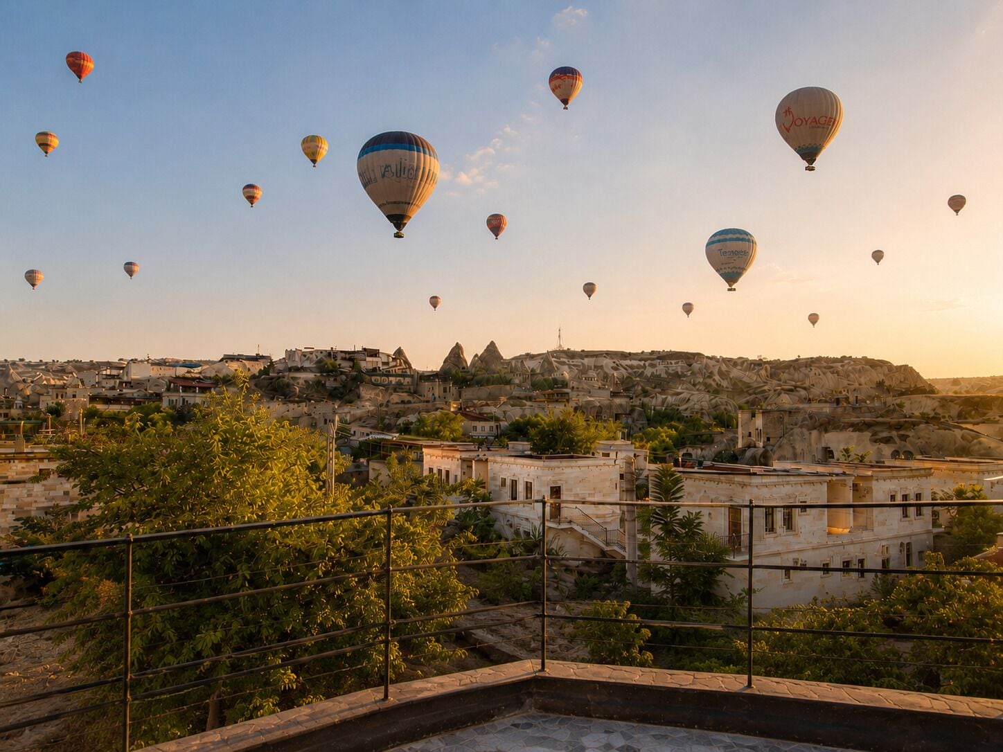Un paisaje urbano con múltiples globos aerostáticos en el cielo, edificios de arquitectura distintiva y una vista despejada del paisaje circundante.
