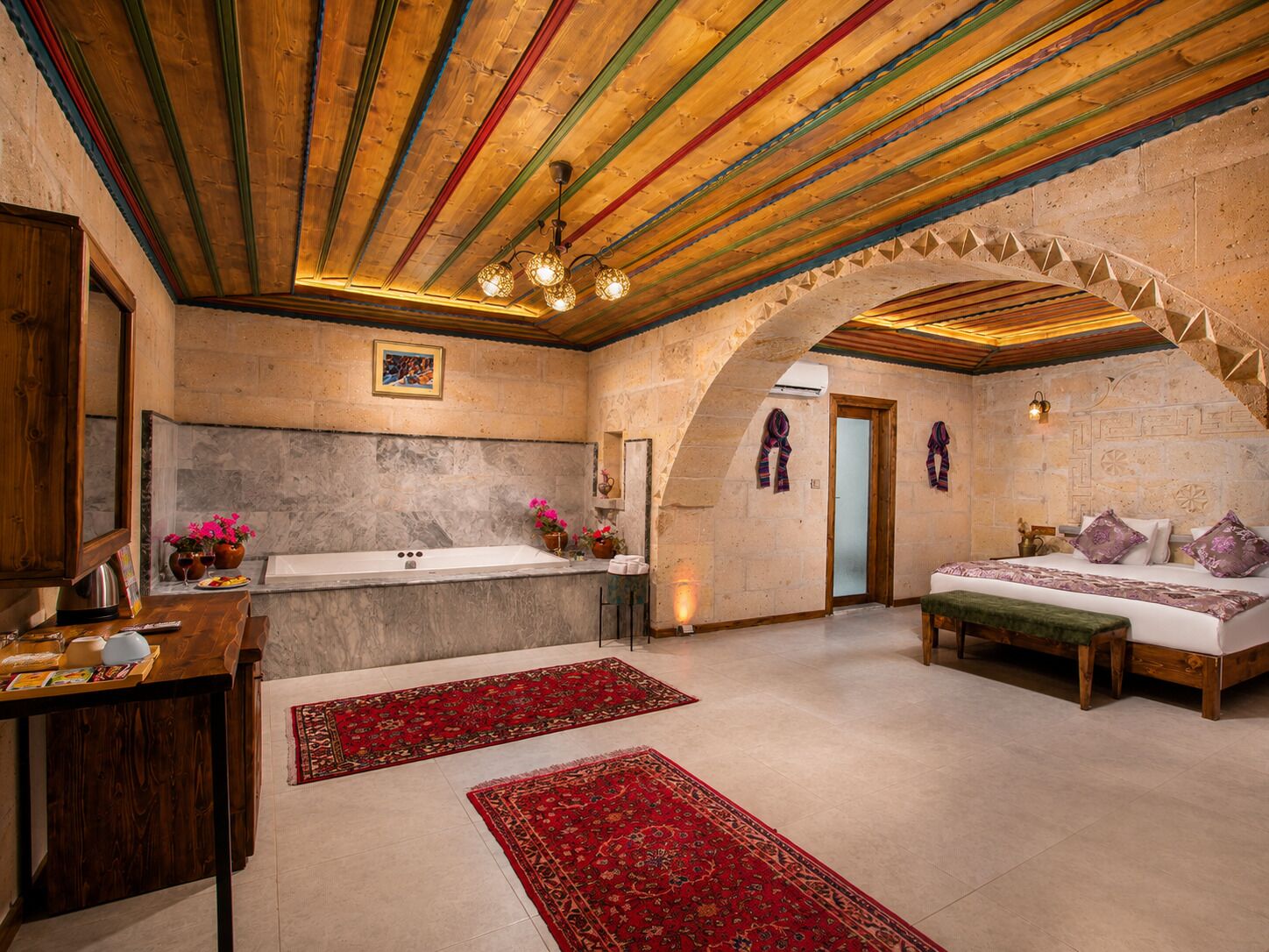 A room with a stone wall, wooden ceiling beams, a bed, a bench, a sink, and a red rug.