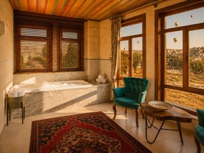 Une pièce avec une baignoire, un fauteuil vert, une petite table avec un bol, et une vue sur un paysage à travers de grandes fenêtres.