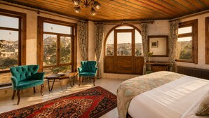 A bedroom with a large bed, two armchairs, a wooden desk, and a view of a mountainous landscape through large windows. - Premium Cappadocia Hotel (Nevsehir)