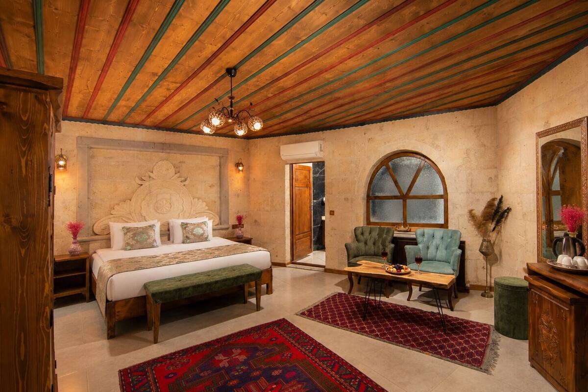 A bedroom with a large bed, a wooden ceiling, a stone wall, a green armchair, a small table with drinks, and a mirror.