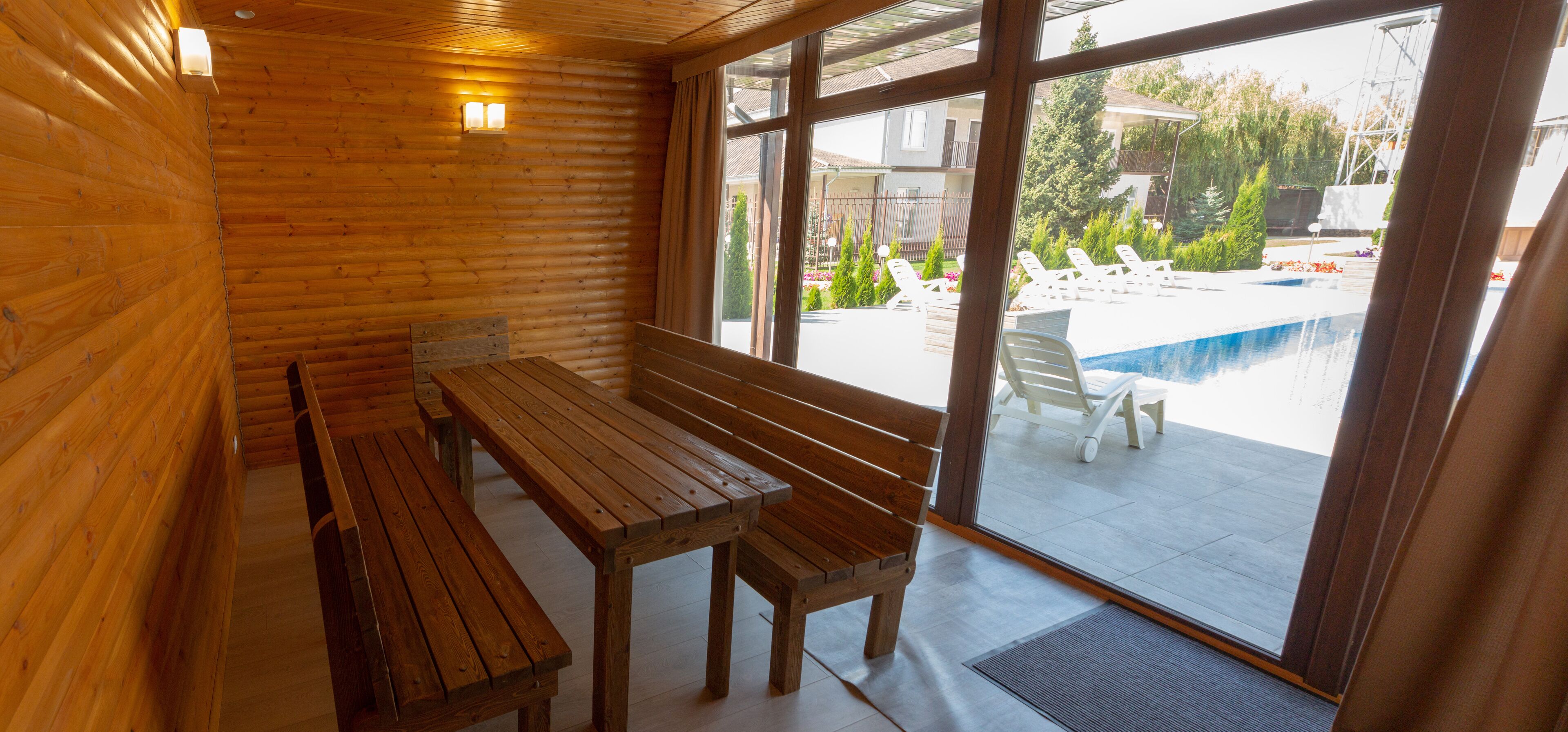 A wooden interior with a bench and table, large windows overlooking a pool area.