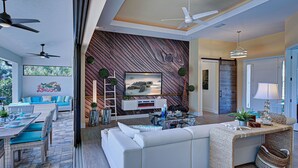 A modern living room with a white sofa, a glass coffee table, and a TV on a wooden wall. There is a dining area with a table and chairs, and a ceiling fan. - Baby SeaTurtle | Pool | Sunshelf | Tiki Bar | Office (Rotonda West)