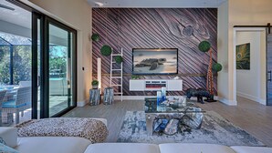 A modern living room with a large flat-screen TV, a glass coffee table, and a sofa. There is a wooden wall with a unique pattern and a ladder. - Baby SeaTurtle | Pool | Sunshelf | Tiki Bar | Office (Rotonda West)