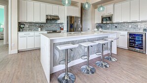 A modern kitchen with white cabinets, a central island with bar stools, a built-in wine cooler, and a backsplash with a stone pattern. - Azure Paradise | Canal View | Dock | 2 Masters | Spa (Port Charlotte)