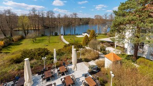 A lakeside patio with outdoor seating and umbrellas, surrounded by trees and a view of the lake. - Residenz Seehotel Berlin Brandenburg (Mittenwalde)