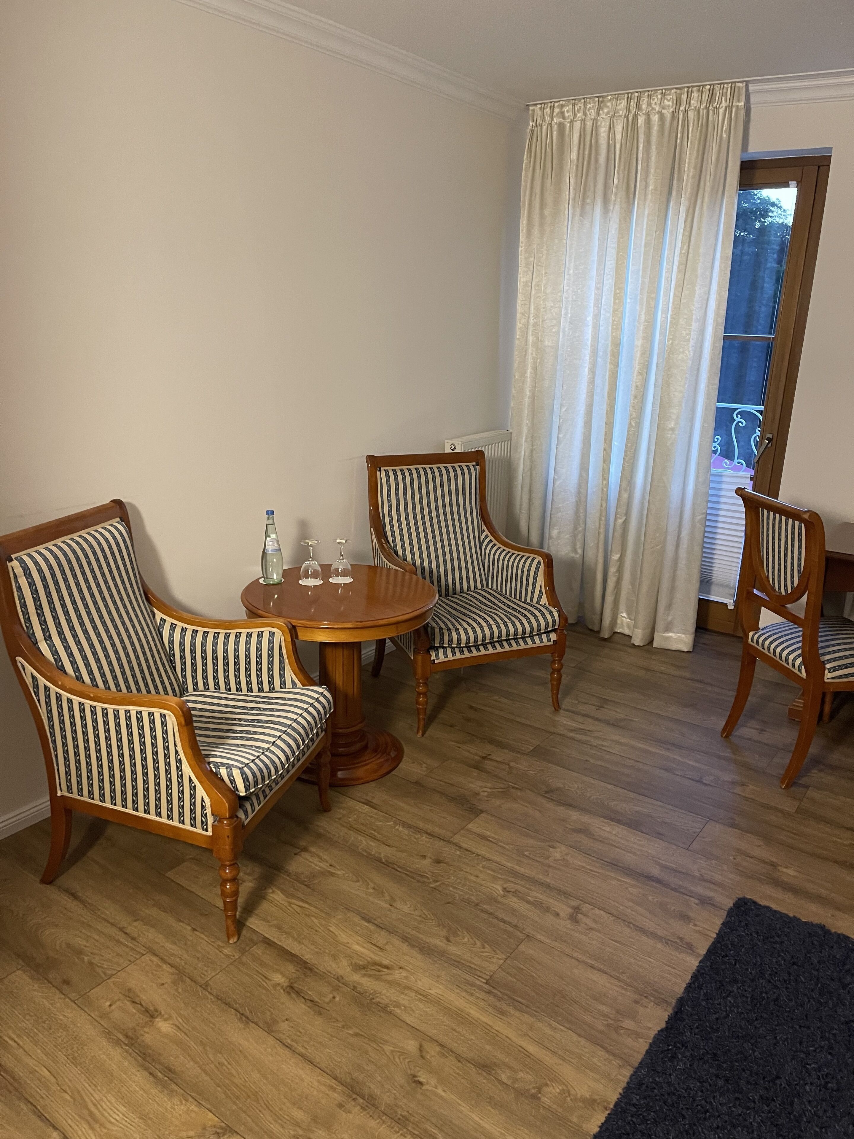 A room with a wooden floor, two striped armchairs, a small round table with a bottle and glasses, and a window with curtains.