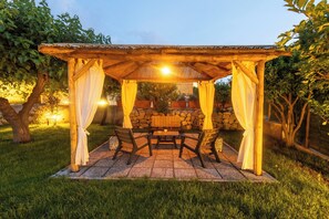 Un belvédère en bois, adossé à un mur de pierres, avec une table et des chaises, le tout entouré de verdure.