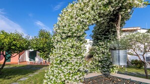 Ein Garten mit einem blühenden Baum, einem Metallpflanzgefäß und einer Terrasse mit Sitzgelegenheiten.