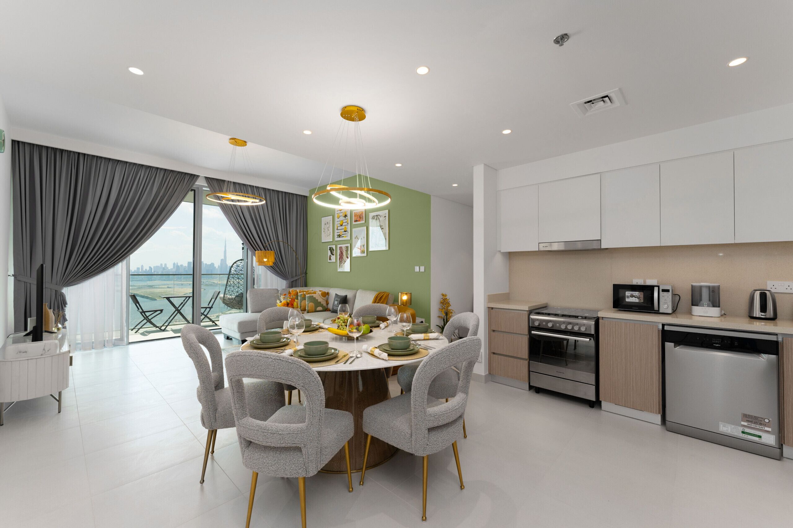 A modern kitchen with stainless steel appliances, a dining area with a round table and grey chairs, and a view of the city through large windows.