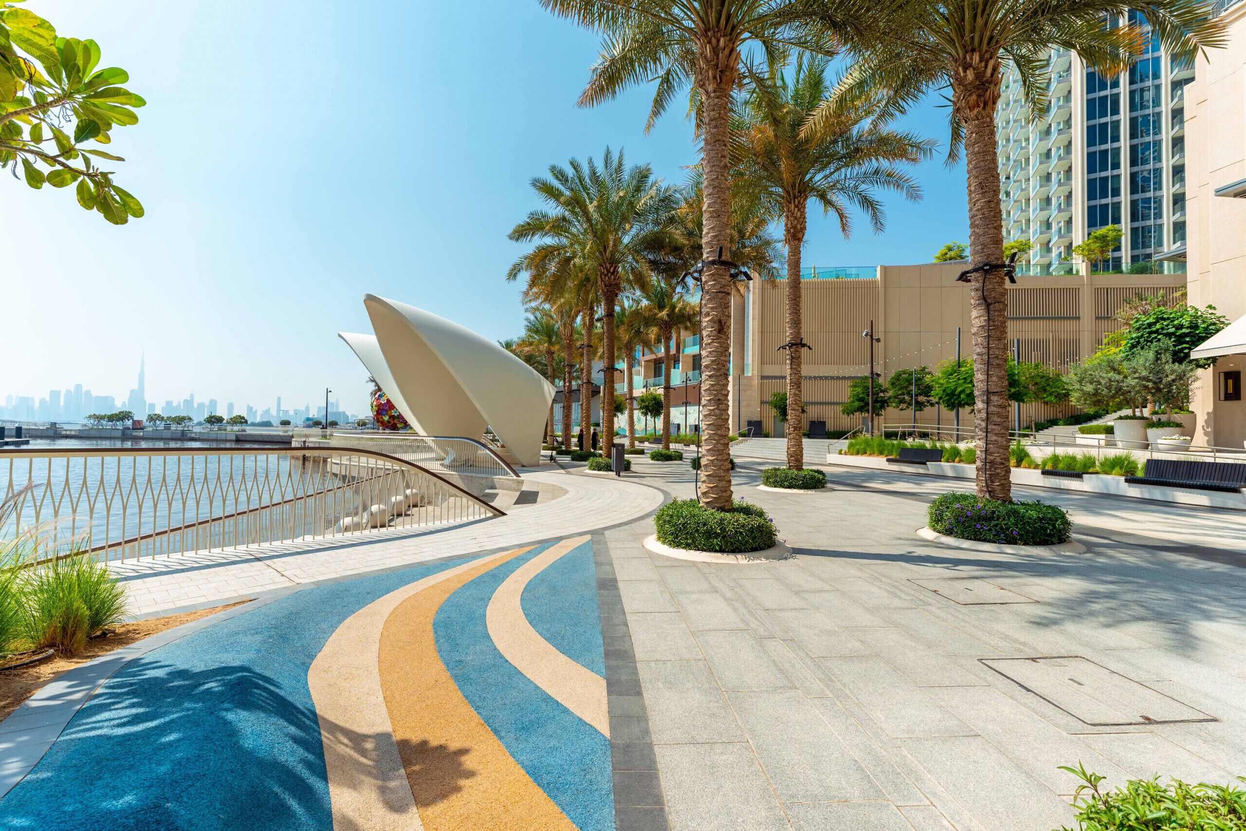 A modern outdoor area with a curved pathway, palm trees, and a waterfront view.