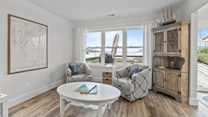 Een woonkamer met uitzicht op de zee, een witte tafel, twee gestreepte banken, een houten kast en een ingelijste kaart aan de muur.