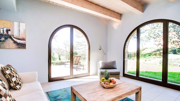 Ein geräumiges Wohnzimmer mit einem Holztisch, einem weißen Sofa und einem Korbstuhl. Große Bogenfenster bieten einen Blick ins Freie.