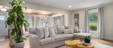 A modern living room with a beige sectional sofa, a large potted plant, a round wooden coffee table, and a view of the outdoors through large windows.