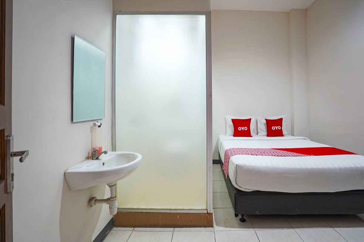 Habitación de hotel con un lavamanos blanco, un espejo, una cama con almohadas rojas y una colcha blanca.