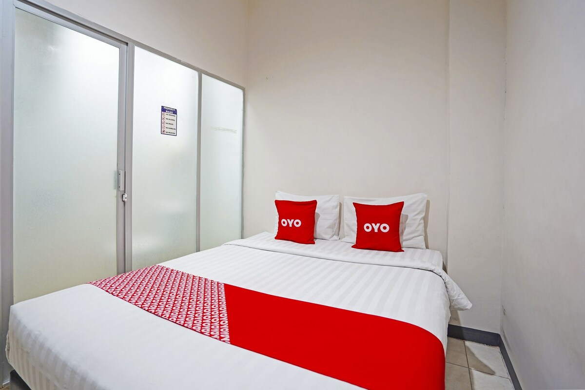Habitación de hotel con cama, dos almohadas rojas con el texto "OYO", una colcha blanca y roja, y una puerta corredera con un cartel.