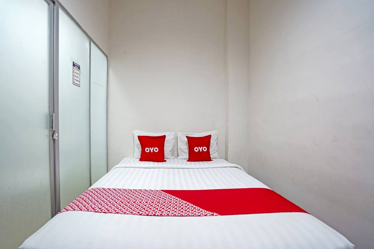 Una habitación de hotel con una cama, dos almohadas con la palabra “OYO” y una manta roja y blanca con estampado, junto a una pared blanca.