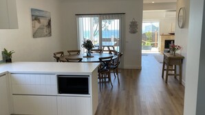 Une cuisine moderne avec un îlot central, une table à manger en bois et des chaises. De grandes fenêtres offrent une vue sur la mer.