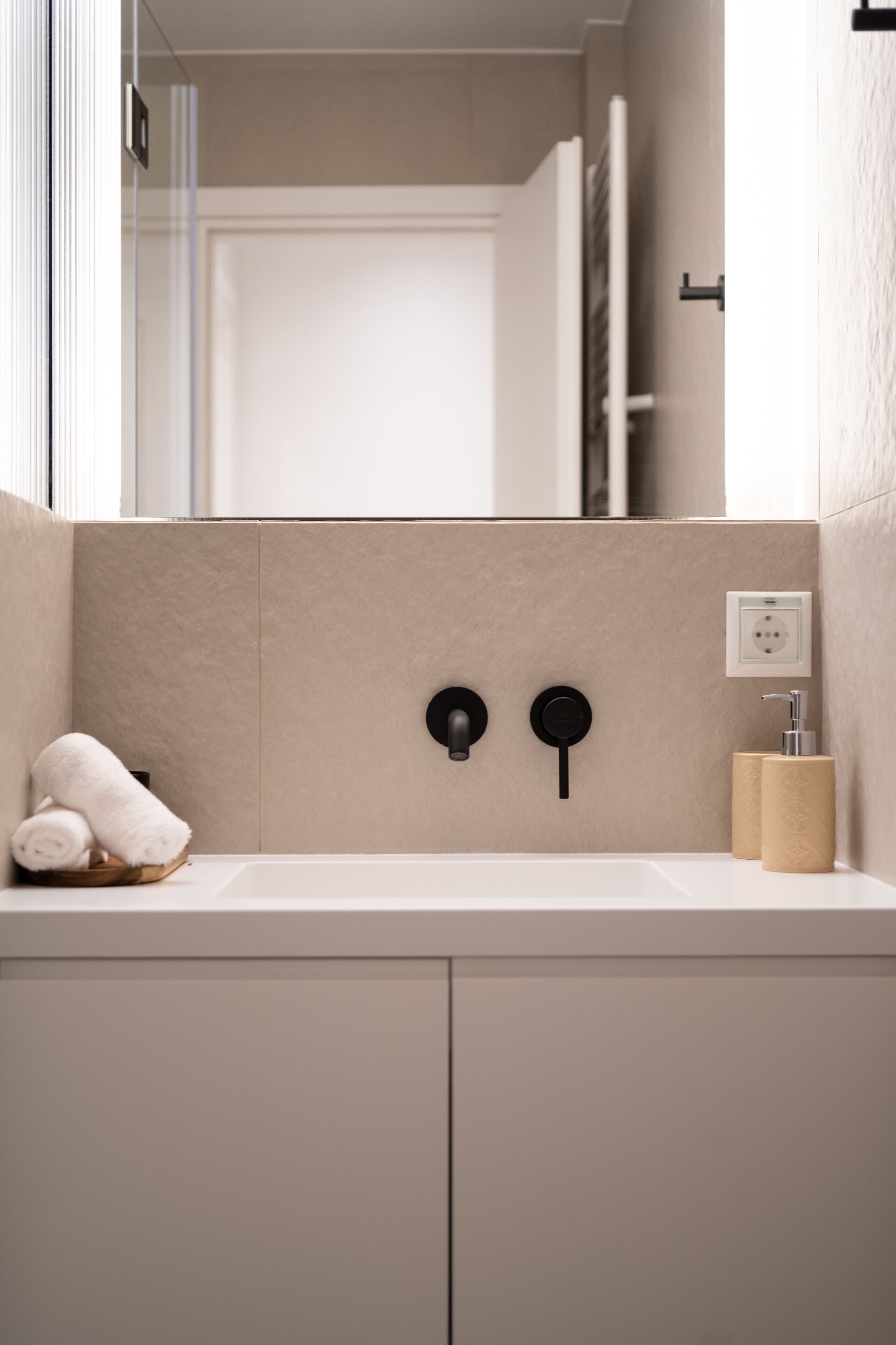 Une salle de bain avec un mur recouvert de carreaux beiges, un lavabo blanc, un porte-serviettes et un distributeur de savon.