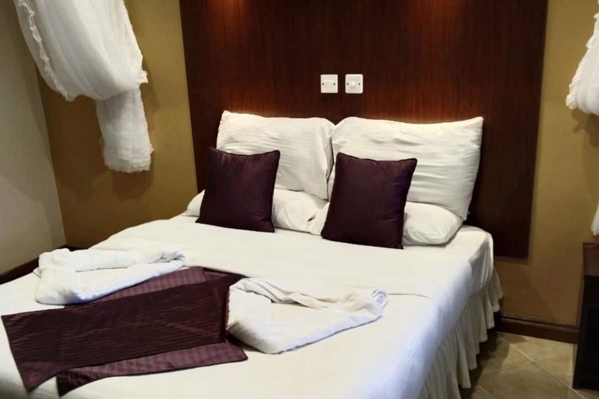 A neatly made bed with white linens and two purple pillows, a wooden headboard, and a white curtain on the left.