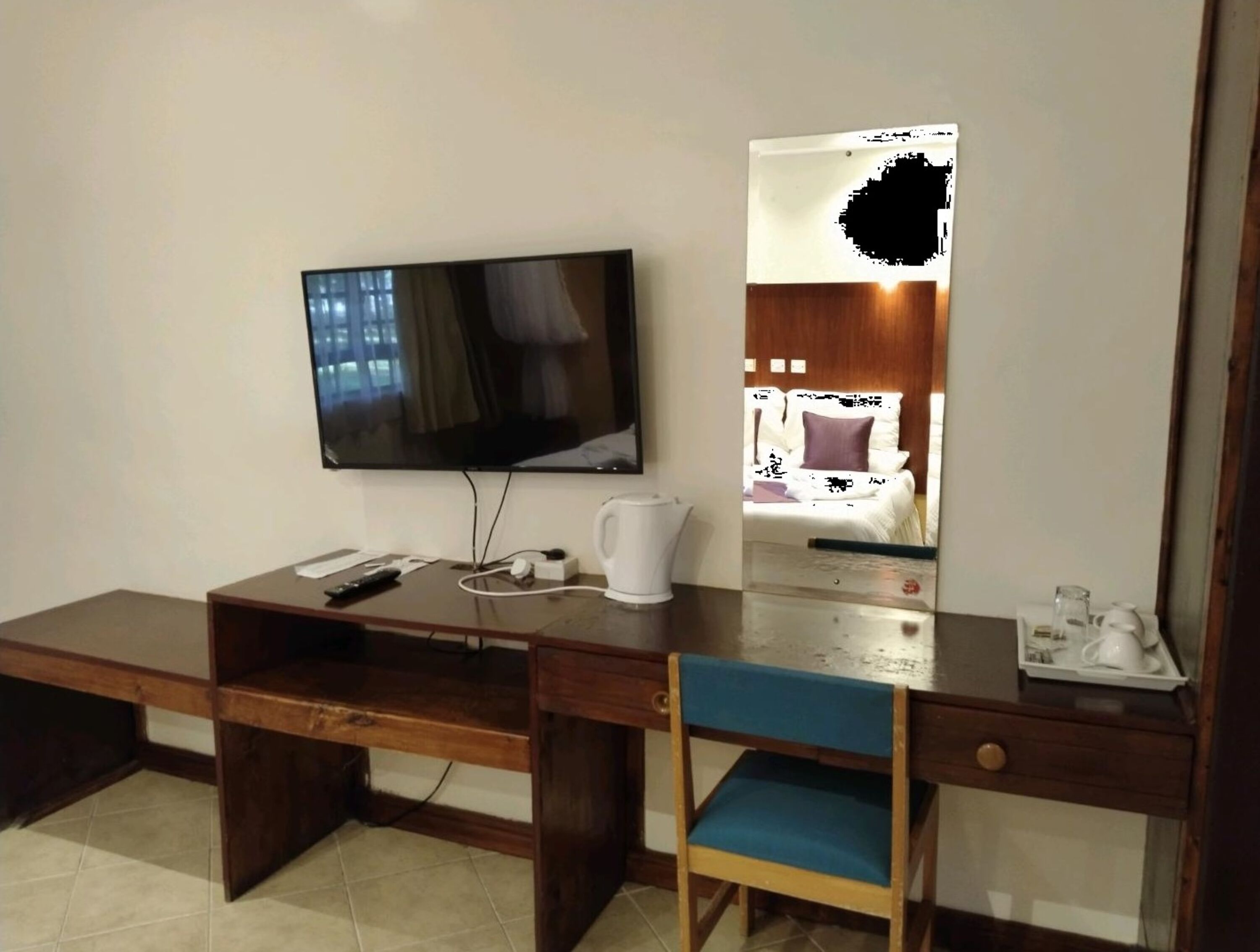 A hotel room with a wooden desk, a television mounted on the wall, a bed with a wooden headboard, and a small table with a kettle and cups.