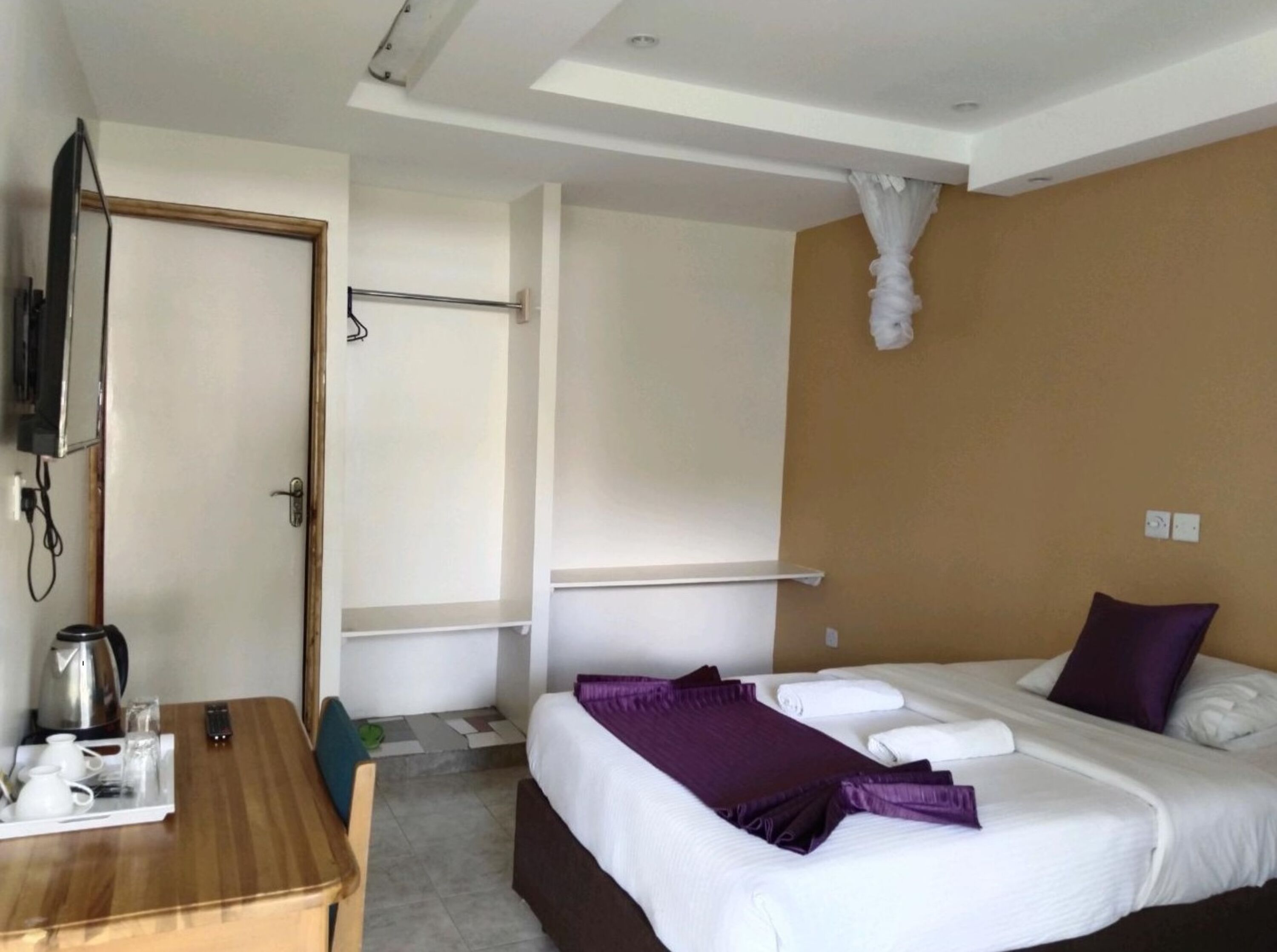 Una habitación de hotel con cama, escritorio con hervidor y bandeja de desayuno, televisión montada en la pared y cortina de ducha.