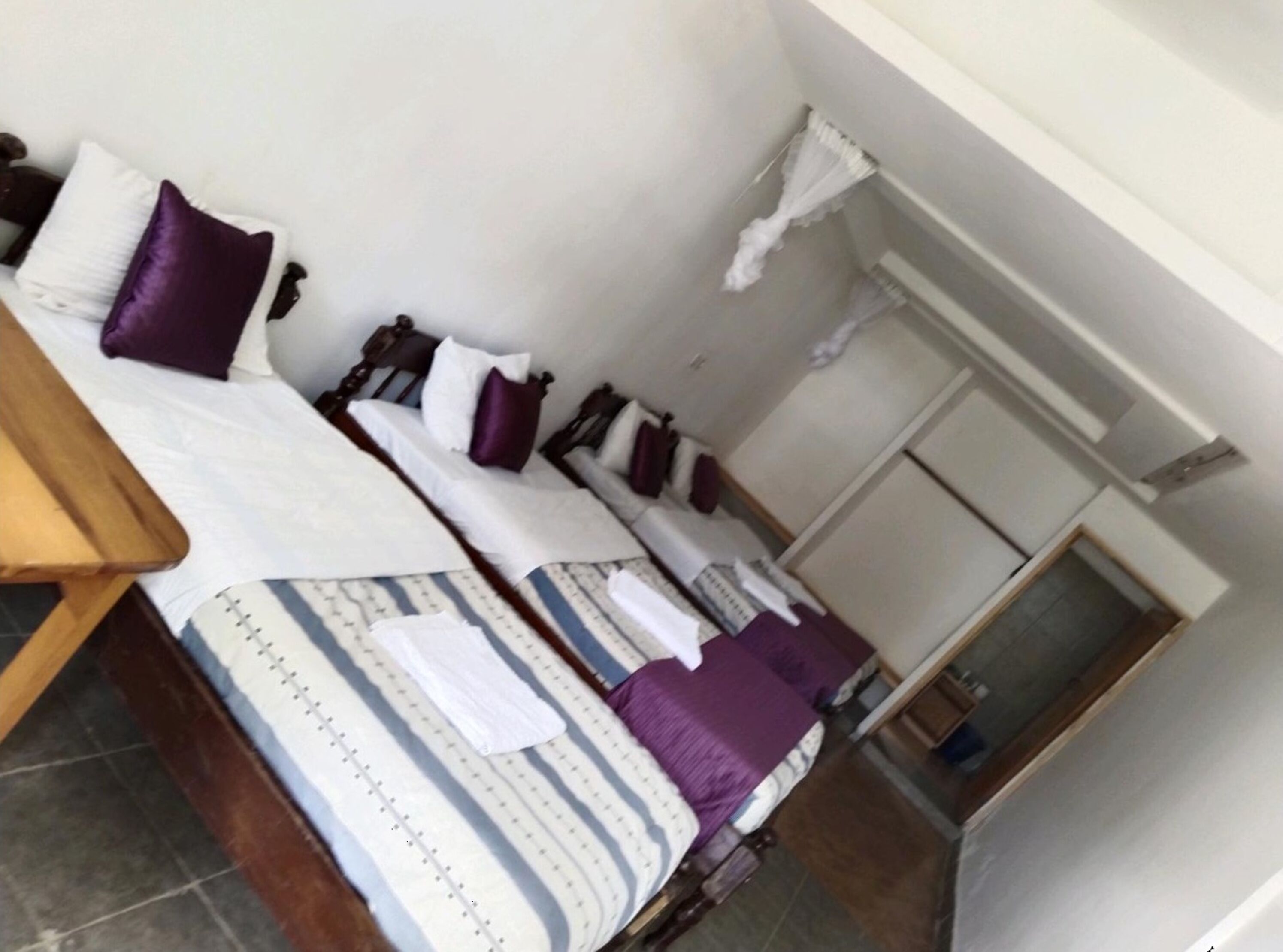 Una habitación con dos camas, cada una con estructura de madera y ropa de cama blanca. Se aprecia una mesita pequeña a la izquierda y una puerta a la derecha.