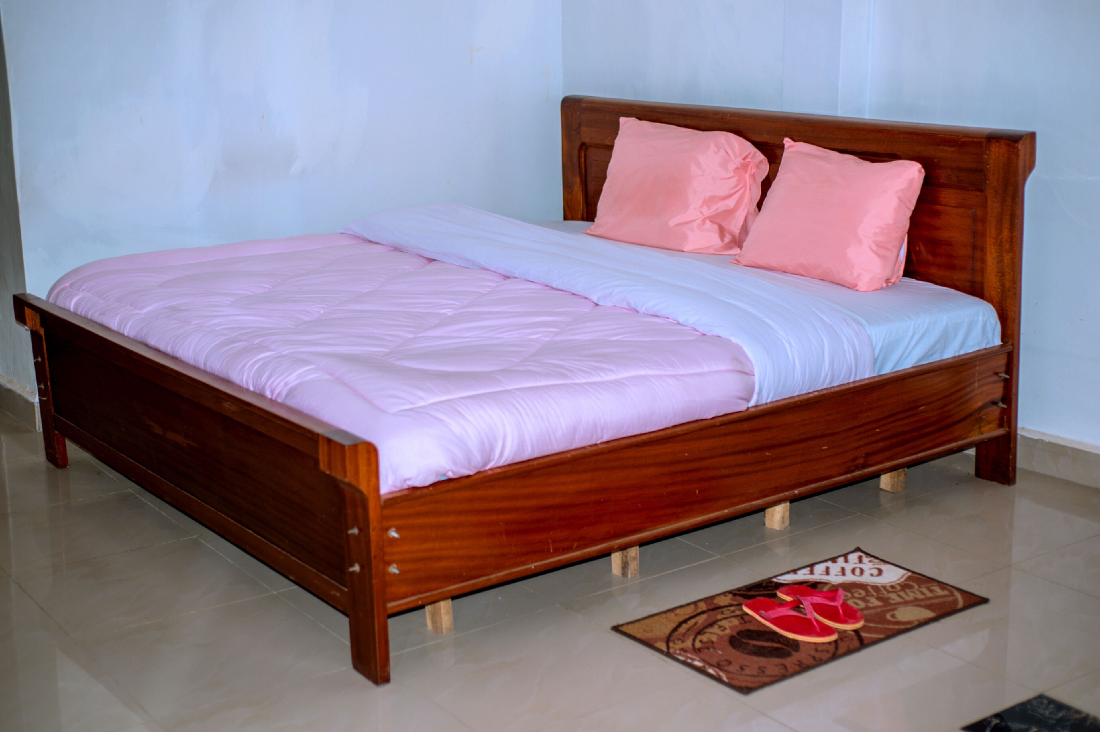 A wooden bed with a pink and white bedspread, two pink pillows, and a doormat with slippers.