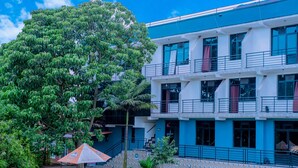 Un edificio de varios pisos con fachada azul y blanca, una escalera que lleva a los pisos superiores y un pequeño jardín con un banco y una sombrilla.