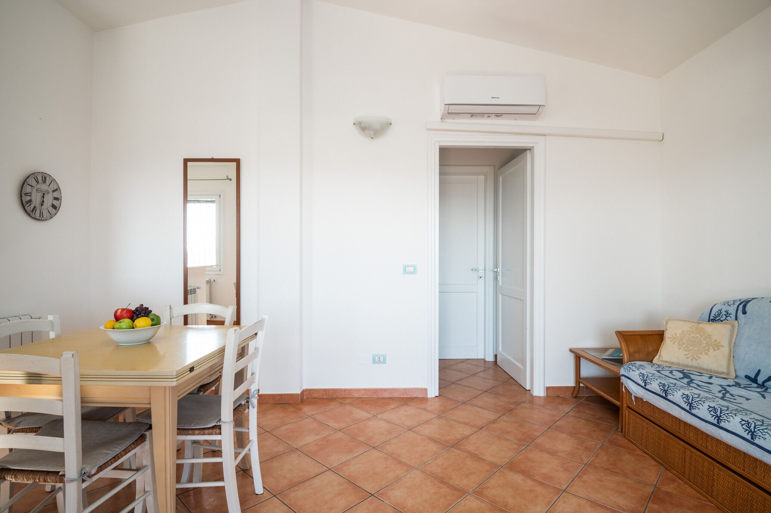 A room with a dining table, chairs, a fruit bowl, a sofa, a mirror, and an air conditioning unit.
