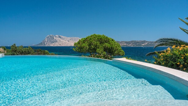 Uma piscina infinita azul cristalina com vista para o mar e uma montanha ao fundo.