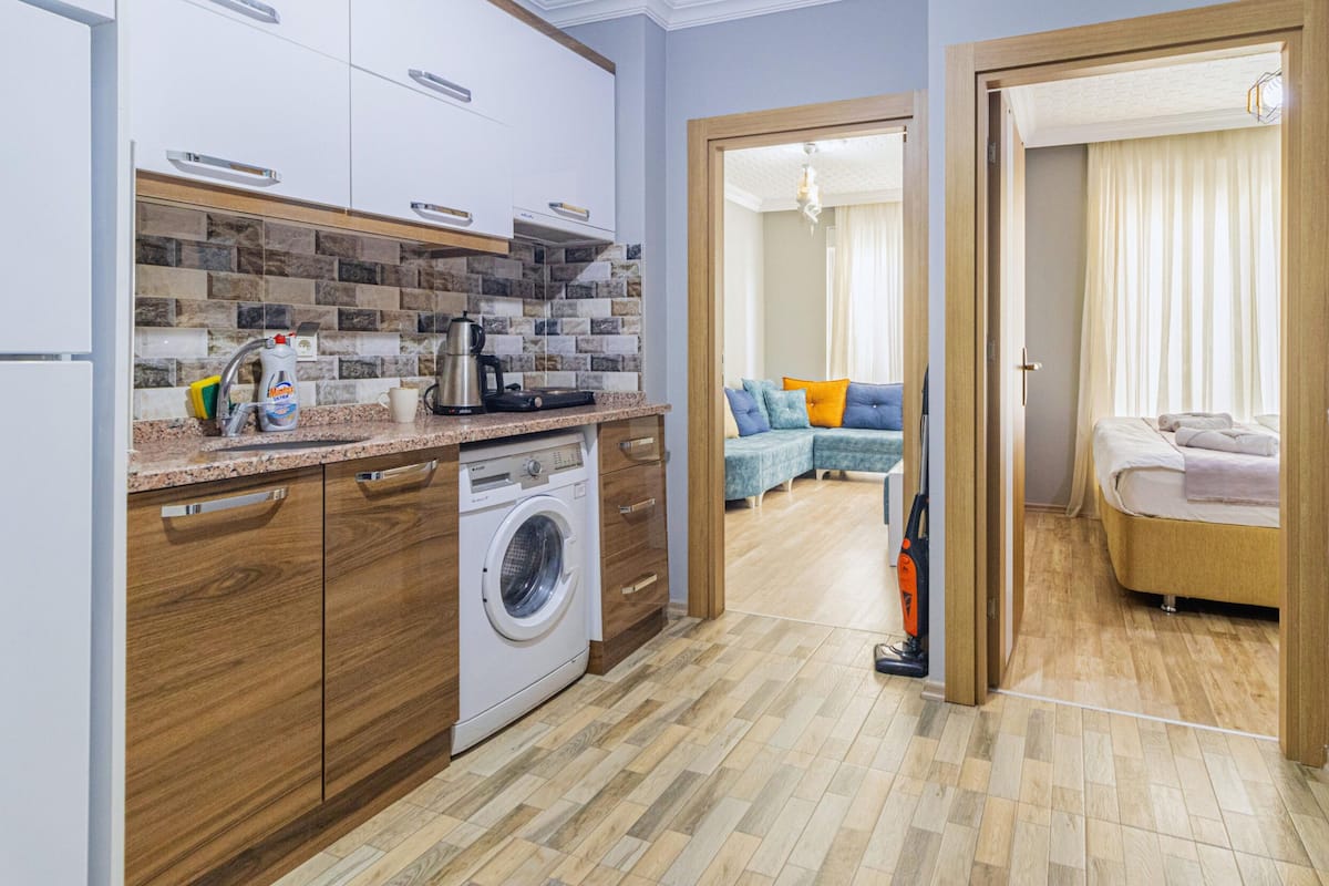 A compact kitchen with a washing machine, a coffee maker, and a sink. A living area with a sofa and a bed is visible through an open door.