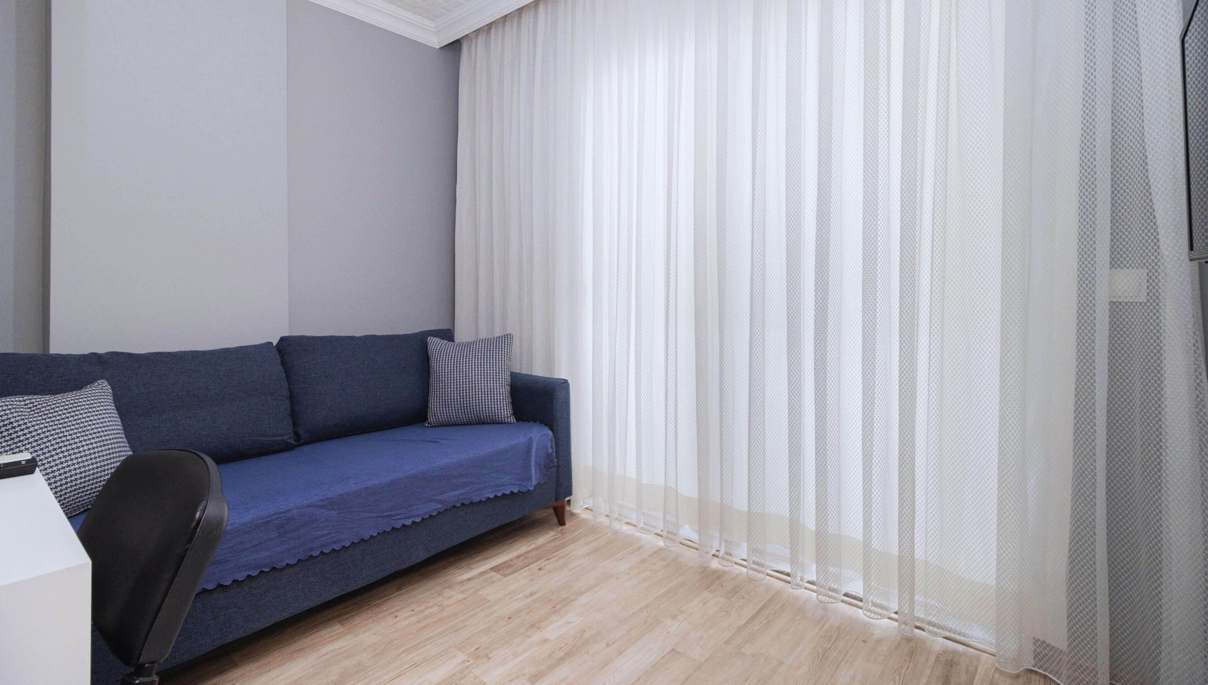 Ein Zimmer mit einem blauen Sofa, einem schwarzen Sessel und durchsichtigen weißen Vorhängen.