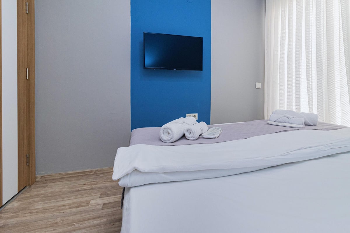 A hotel room with a bed, a flat-screen TV mounted on the wall, and a window with sheer curtains.