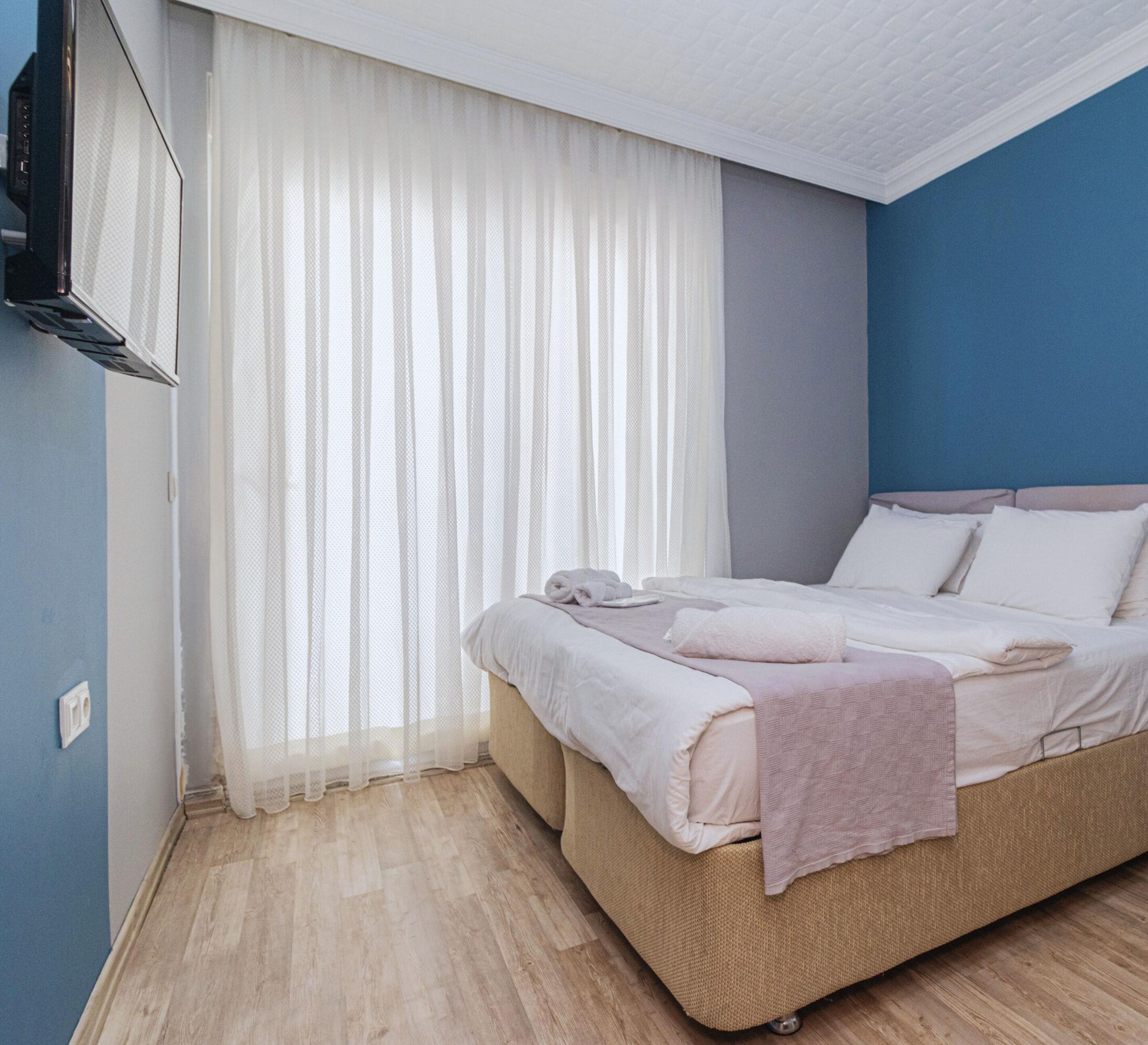 A bedroom with a bed, a TV mounted on the wall, and a window with sheer curtains.